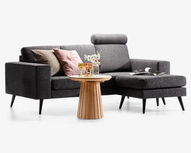 Moderne L-formet sofa med mørkegråt stofbetræk, slanke skrå sorte ben og høj ryg med justerbar nakkestøtte. Sædeområdet har tre pyntepuder i beige, rosa og hvid. Foran står et rundt sofabord i træ med lamelbund, hvorpå der står en vase med hvide blomster og flere farvede glas. Et åbent magasin ligger på chaiselongen og giver et afslappet udtryk.