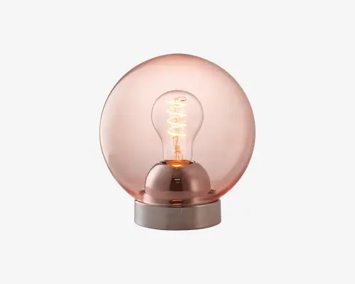 Moderne bordlampe med en stor, rund glaskuppel i en varm rosaguld farve og en synlig glødepære indeni. Lampen har en cylindrisk base i metal, og pæren udsender et blødt, stemningsfuldt lys, der fremhæver den spiralformede glødetråd for et stilfuldt, moderne udtryk.