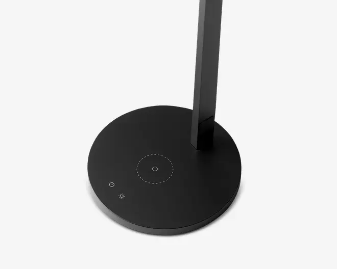 Billedet viser en elegant, rund sort base med mat finish. Basen har en touch-følsom tænd/sluk-knap, et ikon til lysstyrkejustering og et cirkulært område i midten, sandsynligvis til trådløs opladning. Designet er minimalistisk og moderne med diskrete prikkede linjer, der markerer opladningszonen. Den opretstående støtte er fastgjort lidt uden for midten, hvilket giver basen et rent og ryddeligt udseende.