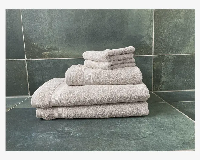 Et pænt stablet sæt lysegrå håndklæder i forskellige størrelser, herunder to små vaskeklude, et håndklæde og to større badehåndklæder. Håndklæderne har en blød, fyldig tekstur og en diskret horisontal kantdetalje nær enderne. De er placeret på en mørkegrøn fliseoverflade, hvilket antyder et badeværelsesmiljø.