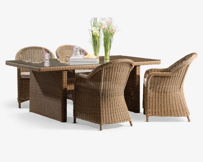 Sættet består af et rektangulært bord med glasplade og fire matchende armstole, alle fremstillet af flettet rattan i en naturlig lysebrun farve. Stolene har buede armlæn og høje rygge for komfort samt hvide sædehynder for ekstra støtte. Bordet er pyntet med en vase med friske blomster og et fad med en kande og glas, hvilket giver et indbydende og stilfuldt udendørs spiseområde.