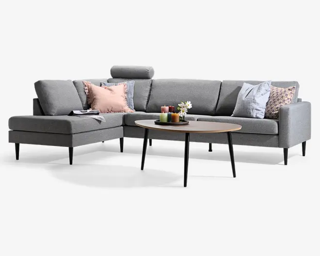 Grå L-formet hjørnesofa med chaiselong til venstre, lave ryghynder, sorte koniske ben og assorterede pyntepuder i rosa, blå og mønstrede stoffer. Et rundt sofabord i træ med sorte ben har stearinlys og en lille blomsterdekoration.