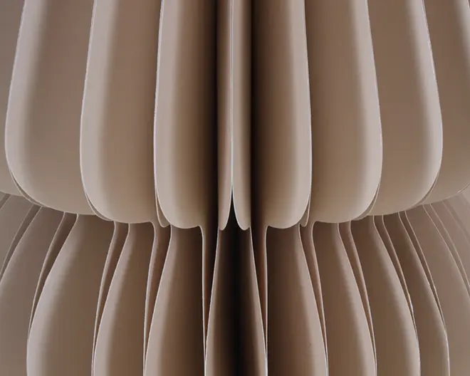 Nærbillede af en struktur lavet af flere lag buet, beige materiale arrangeret i et symmetrisk, lodret mønster. Lagene skaber en blød, skulpturel effekt med afrundede kanter og dybe riller, hvilket giver overfladen et tredimensionelt, tekstureret udseende.