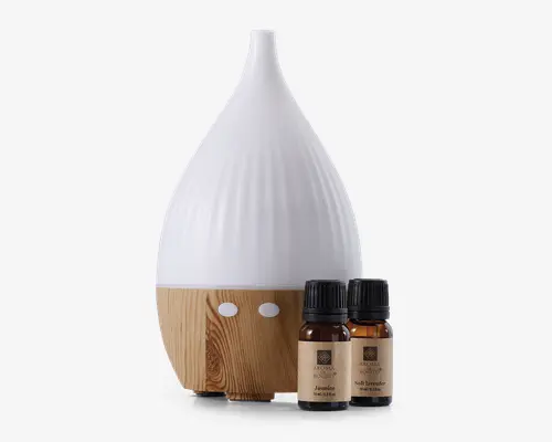 En dråbeformet enhed med mat hvid top og lys træstruktur i bunden, udstyret med to runde hvide knapper. Ved siden af står to små brune glasflasker med sorte låg, mærket 'Jasmine' og 'Soft Lavender'. Designet er moderne og minimalistisk med naturlige trædetaljer og tydelig mærkning på flaskerne.
