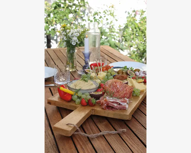 Et træskærebræt er anrettet med forskellige skiver af charcuteri, oste, friske vindruer, jordbær og skiver af peberfrugt. Der er også små skåle med dip eller smørepålæg, og brættet er pyntet med friske krydderurter. Brættet står på et udendørs træbord, hvor der også er en vase med vilde blomster, en lysestage med et hvidt lys og en klar glasflaske med vand i baggrunden, hvilket skaber en rustik og indbydende præsentation.