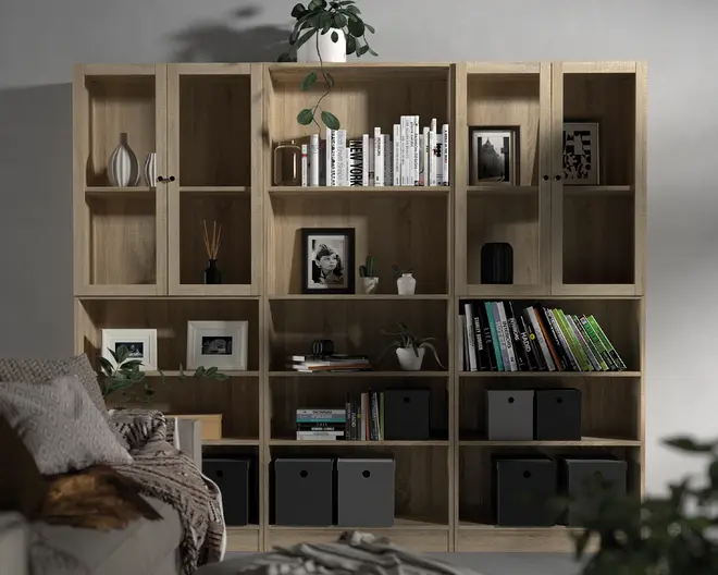 Høj reol i lyst træ med en kombination af åbne hylder og sektioner med glaslåger. Hylderne indeholder forskellige genstande såsom bøger arrangeret både lodret og vandret, dekorative vaser, indrammede fotografier, små potteplanter og sorte opbevaringskasser. Reolen er indrettet i en moderne, minimalistisk stil og står op ad en lys væg.