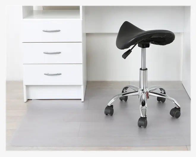 En gennemsigtig, rektangulær gulvmåtte placeret under en kontorstol med hjul, krom base og sort sadelformet sæde. Måtten beskytter det lyse trægulv mod ridser og slid fra stolens hjul. Måtten har en glat overflade og passer pænt foran et hvidt skrivebord med tre skuffer.