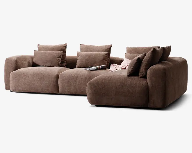 En stor, moderne hjørnesofa med chaiselong til højre, betrukket med et tekstureret brunt stof. Sofaen har brede, dybe sæder og tykke, afrundede armlæn for ekstra komfort. Den leveres med seks matchende ryghynder og to mindre pyntepuder, som giver god støtte. Et magasin og et let tæppe ligger afslappet på chaiselongen, hvilket skaber en hyggelig og indbydende stemning.