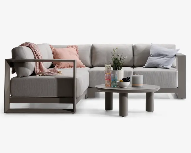 Moderne L-formet sofa med lysegråt betræk og robuste, vinkelrette armlæn i metal. Sofaen har bløde sæde- og ryghynder og er pyntet med et lyserødt tæppe, en lyserød pyntepude og en lyseblå pude for ekstra komfort. Foran står et rundt, lavt sofabord med en potteplante, et sæt farverige glas, et hvidt lys og en skål med bær, hvilket skaber en hyggelig og indbydende stemning.