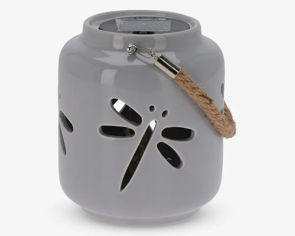 En rund, blank keramiklanterne med udskåret guldsmed-design på siden. Lanternen er lysegrå og har et robust rebhåndtag, der er fastgjort med metalbeslag. Øverst på lanternen sidder et solpanel, hvilket viser, at den er solcelledrevet til belysning.