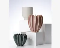 Billedet viser tre keramiske urtepotter med forskellige designs. Den bagerste potte er hvid, cylindrisk og har lodrette riller. Den midterste potte er lyserød med blank overflade og en rund, bølget form, der minder om blomsterblade eller et græskar. Den forreste potte er mørkegrøn, også blank, og har en lignende bølget, organisk form. Alle potterne har et moderne, dekorativt udtryk og er placeret på hvide, geometriske sokler.