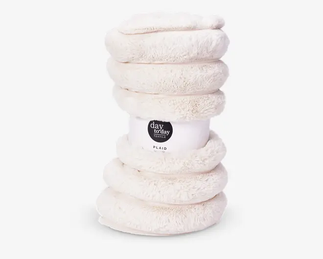 Et sammenrullet, plysbetrukket tæppe i cremefarve med en blød og fluffy tekstur. Tæppet er holdt sammen af et papbånd omkring midten, som viser mærkenavn og produktinformation. Materialet ser tykt og hyggeligt ud, ideelt til varme og komfort.