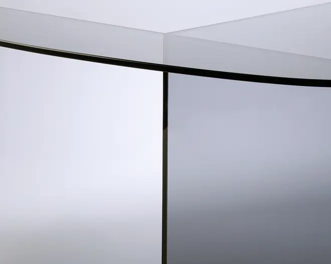Nærbillede af et møbel med et slankt, moderne design. Overfladen og støttepanelet er lavet af reflekterende, tonet glas med rene, skarpe kanter. Glasset har en let grålig tone, og konstruktionen fremhæver minimalisme og elegance.