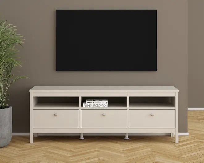Et moderne, lysebeige TV-bord med et enkelt, minimalistisk design. Det har tre rummelige skuffer med runde greb nederst og tre åbne hylder ovenover. En af hylderne indeholder en stak bøger. Bordet står på fire solide ben og har en glat, flad top, der egner sig til at bære et fjernsyn eller pyntegenstande. Baggrunden består af en taupefarvet væg, et stort sort rektangulært panel over bordet og en potteplante til venstre.