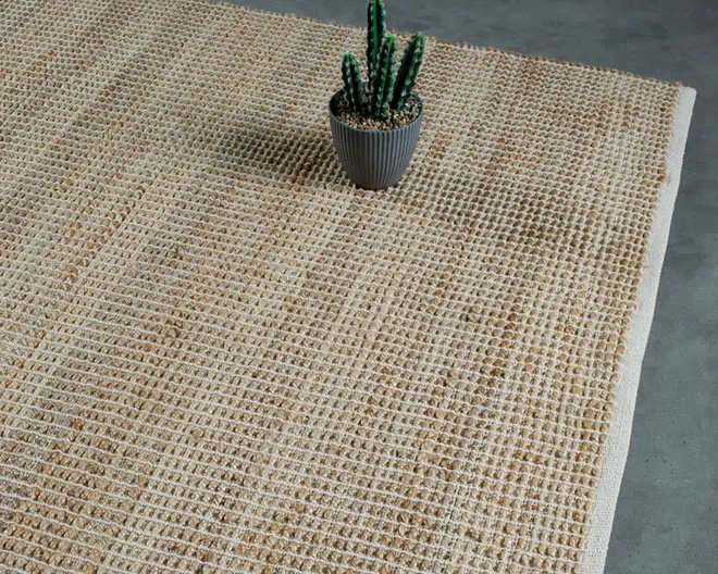 Billedet viser et nærbillede af et vævet tæppe i en naturlig, beige farve. Tæppet har en tekstureret, gitterlignende overflade, der er skabt af sammenflettede tykke og tynde fibre, hvilket giver det et rustikt og organisk udtryk. Kanterne er pænt afsluttede, og overfladen ser slidstærk og robust ud. En lille kaktus i potte står på tæppet og fremhæver dets neutrale tone og alsidige stil.