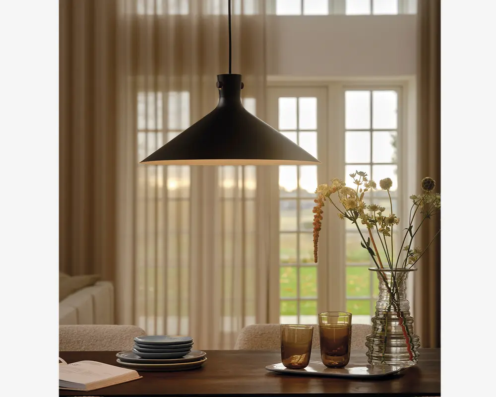 En mat sort pendellampe med en bred, konisk skærm hænger over et spisebord. Lampen har et glat, minimalistisk design med en let udadgående kant, der giver fokuseret lys nedad. På bordet nedenunder står stabelde keramiktallerkener, to farvede glas og en klar glasvase med en blanding af tørrede og friske blomster. I baggrunden ses lyse gardiner og store vinduer, som lader naturligt lys blødt oplyse rummet.