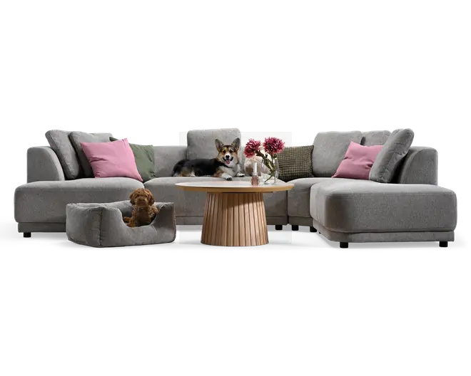 Lysegrå modulopbygget hjørnesofa med afrundede armlæn og dybe sædehynder, stylet med rosa og grønne pyntepuder, et rundt sofabord i træ og en matchende kæledyrsseng.