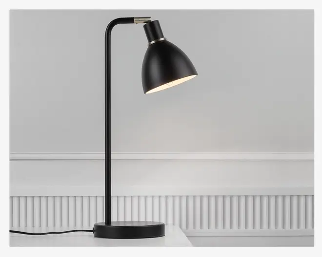 En moderne bordlampe med mat sort finish, rund fod, lige lodret stang og en vippet, kuppelformet skærm. Lampen har et minimalistisk design med synlig ledning og en metal-detalje ved samlingen mellem skærm og stang. Skærmen kan justeres, så lysets retning kan ændres.