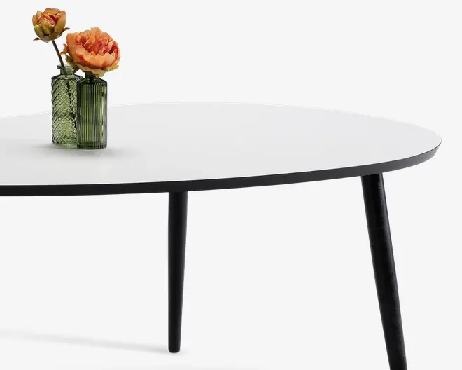 Billedet viser et rundt bord med en glat, hvid bordplade og slanke, sorte ben. På bordet står to dekorative grønne glasvaser med mønster, hver med orange og gule kunstige blomster. Bordet har et minimalistisk og moderne design.