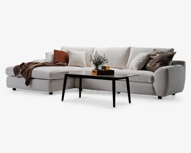 En moderne L-formet sofa betrukket med lysebeige stof, med brede armlæn og bløde ryghynder. Sofaen er pyntet med flere pyntepuder i forskellige farver og mønstre, herunder hvide, brune og geometriske designs. Et blødt brunt tæppe er lagt over den ene side. Foran sofaen står et elegant sort sofabord med tynde ben, hvorpå der står en lille dekorativ plante og en træbakke med kopper.
