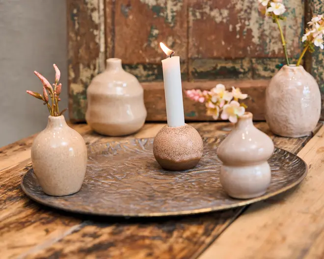 En dekorativ opstilling med små, runde og unikt formede keramikvaser i bløde beige og cremefarver, nogle med fine kunstige blomster. I midten står en matchende keramikstage med et tændt hvidt kronelys. Alle genstande er placeret på et stort, tekstureret metalfad oven på et rustikt træbord, hvilket skaber et hyggeligt og elegant udtryk.