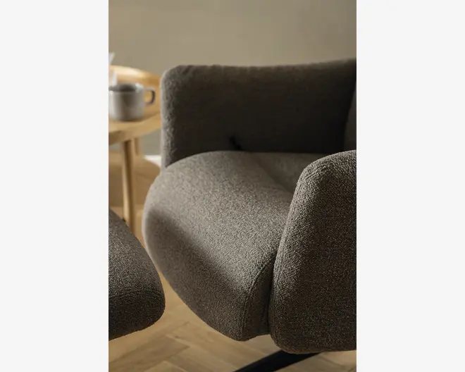 Nærbillede af en moderne lænestol med blødt, tekstureret mørkegråt betræk. Stolen har tykke, afrundede armlæn og et bredt, polstret sæde for ekstra komfort. Stoffet har et diskret, vævet udseende. I baggrunden ses et lyst træbord med en kop og en kande, og gulvet er af lyst træ.