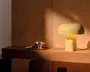 Lille gul lampe i mat finish med rund svampeformet skærm, cylindrisk fod og synlig ledning, der kaster et varmt lys på en træhylde.