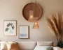 Et hyggeligt hjørne af en stue med en sofa i neutrale farver og bløde puder i beige og sandfarver. Over sofaen hænger to pendellamper med flettede rattan-skærme i forskellige højder, som giver et varmt lys. Bag lamperne er der en stor, rund vægdekoration i flet, der tilføjer tekstur. På væggen hænger også to indrammede billeder: et med en botanisk illustration og et med et minimalistisk havmotiv. Til højre står høje pampasgræs i en vase, hvilket giver rummet et naturligt og luftigt udtryk.