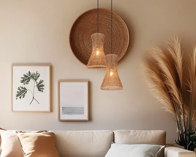 Et hyggeligt hjørne af en stue med en sofa i neutrale farver og bløde puder i beige og sandfarver. Over sofaen hænger to pendellamper med flettede rattan-skærme i forskellige højder, som giver et varmt lys. Bag lamperne er der en stor, rund vægdekoration i flet, der tilføjer tekstur. På væggen hænger også to indrammede billeder: et med en botanisk illustration og et med et minimalistisk havmotiv. Til højre står høje pampasgræs i en vase, hvilket giver rummet et naturligt og luftigt udtryk.