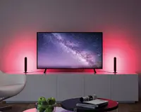 Et moderne underholdningssetup med et fladskærms-tv placeret på et hvidt skab. Tv'et viser en stjerneklar nattehimmel med Mælkevejen. På hver side af tv'et står der to lodrette LED-lysstave, som udsender et blødt lyserødt-rødt skær, der oplyser væggen bagved. Skabet er minimalistisk med rene linjer, og i forgrunden står et rundt sofabord med en skål grønne æbler og en stak magasiner. En hvid, buet stol er delvist synlig til venstre.