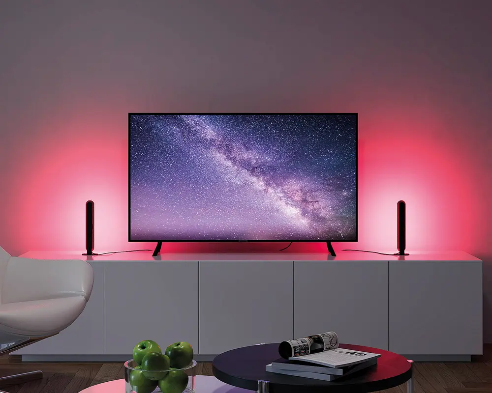 Et moderne underholdningssetup med et fladskærms-tv placeret på et hvidt skab. Tv'et viser en stjerneklar nattehimmel med Mælkevejen. På hver side af tv'et står der to lodrette LED-lysstave, som udsender et blødt lyserødt-rødt skær, der oplyser væggen bagved. Skabet er minimalistisk med rene linjer, og i forgrunden står et rundt sofabord med en skål grønne æbler og en stak magasiner. En hvid, buet stol er delvist synlig til venstre.