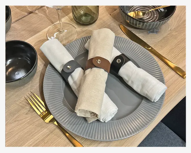 Tre stofservietter rullet sammen og holdt på plads med servietringe i sort og brun læder, hver med en metallisk trykknap. Servietterne ligger på en rillet, lysegrå keramik tallerken. Borddækningen inkluderer bestik i guldfarve, en sort skål, en rillet grå skål med en guldske, et vinglas og et grønligt glas, alt sammen arrangeret på et lyst træbord.