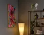 Et højt indrammet abstrakt print med kraftige penselstrøg i pink, blå og gul, vist ved siden af en gulvlampe med varmt lys og en sort metalskænk med planter, bøger og dekorative vaser.