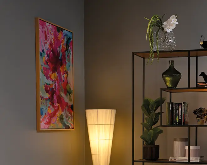 Et højt indrammet abstrakt print med kraftige penselstrøg i pink, blå og gul, vist ved siden af en gulvlampe med varmt lys og en sort metalskænk med planter, bøger og dekorative vaser.