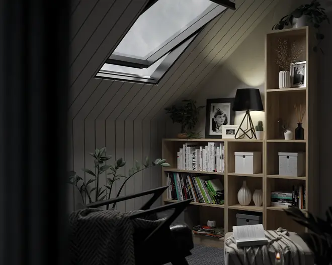 Et moderne reolsystem i træ med naturfinish, der kombinerer åbne hylder og kuber. Hylderne er pænt organiseret med bøger, dekorative hvide opbevaringskasser og minimalistiske vaser. Nogle hylder har indrammede fotos og små potteplanter, mens en sort bordlampe med geometrisk fod står ovenpå og spreder et varmt lys. Reolen står op ad en skrå væg under et ovenlysvindue, hvilket skaber en hyggelig og stilfuld læsekrog.