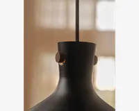 Nærbillede af en lampeskærm i mat sort metal med en glat, buet form. Lampeskærmen har to runde træknapper på hver sin side nær toppen, hvilket giver et varmt og kontrastfuldt udtryk til det moderne design. En stofbetrukket ledning går ud fra midten i toppen til ophæng.