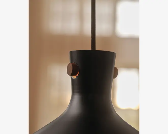 Nærbillede af en lampeskærm i mat sort metal med en glat, buet form. Lampeskærmen har to runde træknapper på hver sin side nær toppen, hvilket giver et varmt og kontrastfuldt udtryk til det moderne design. En stofbetrukket ledning går ud fra midten i toppen til ophæng.