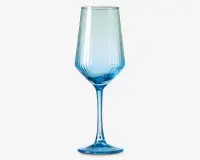 Højt glas med blå nuance, lang stilk og bred, vinklet skål. Skålen har lodrette rillede linjer, der giver tekstur og et dekorativt udtryk. Bunden er rund og solid og matcher glassets blå farve.