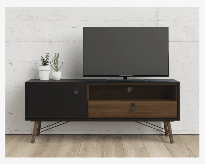Et moderne TV-bord med et minimalistisk design, der har en mat sort overflade kombineret med en skuffe og en åben hylde i naturtræ. Bordet har skrå træben og metalstøtter for stabilitet. Til venstre er der et lukket skab med et lille firkantet greb, mens højre side har en åben hylde over en bred træskuffe med et rektangulært greb. To små potteplanter står ovenpå bordet ved siden af fjernsynet.