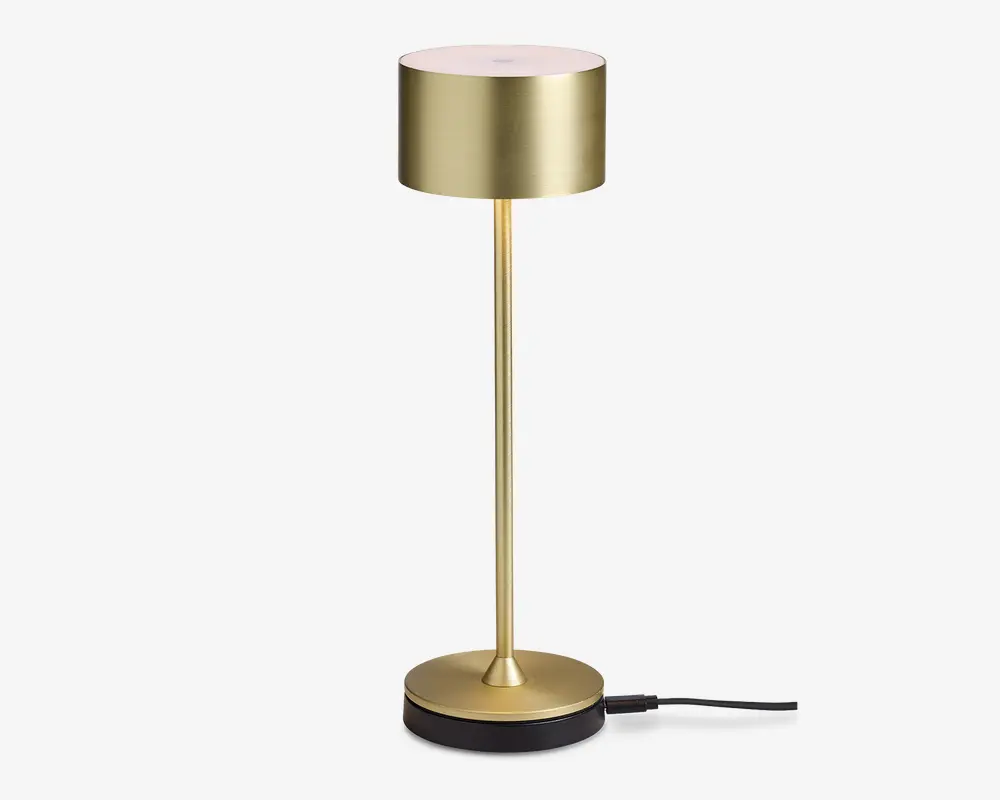 Slank, moderne lampe med børstet guldfinish, cylindrisk lampeskærm, tynd lodret stang og rund, tung fod. Lampen har et minimalistisk design og berøringskontrol på toppen af skærmen.