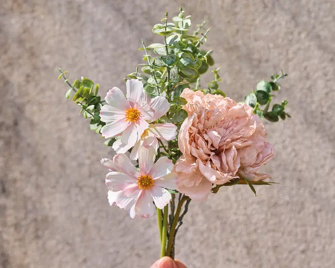 En lille buket med tre lyserøde margueritlignende blomster med gule midter, en stor, ruflet, lyserød pæon og flere stilke med runde, grønne eukalyptusblade. Blomsterne og det grønne er arrangeret sammen og viser en blanding af fine kronblade og tekstureret løv.