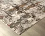 Rektangulært tæppe med et moderne abstrakt mønster i grå, hvid, rustorange og kulfarvede nuancer. Designet har et slidt, tekstureret udseende med uregelmæssige felter og linjer, hvilket giver et moderne og kunstnerisk præg. Tæppet har lav luv og pænt afsluttede kanter, velegnet til moderne indretning.
