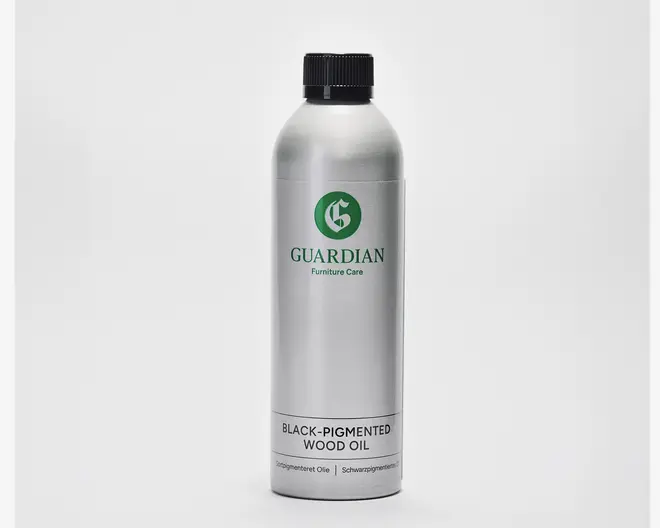 En høj, cylindrisk sølvfarvet metalflaske med sort skruelåg. Etiketten har mærket 'Guardian Furniture Care' i grøn samt et grønt cirkulært logo. Nederst på etiketten står der 'Black-Pigmented Wood Oil' med fed sort skrift, og der er yderligere tekst i mindre skrift nedenunder. Designet er minimalistisk og professionelt.
