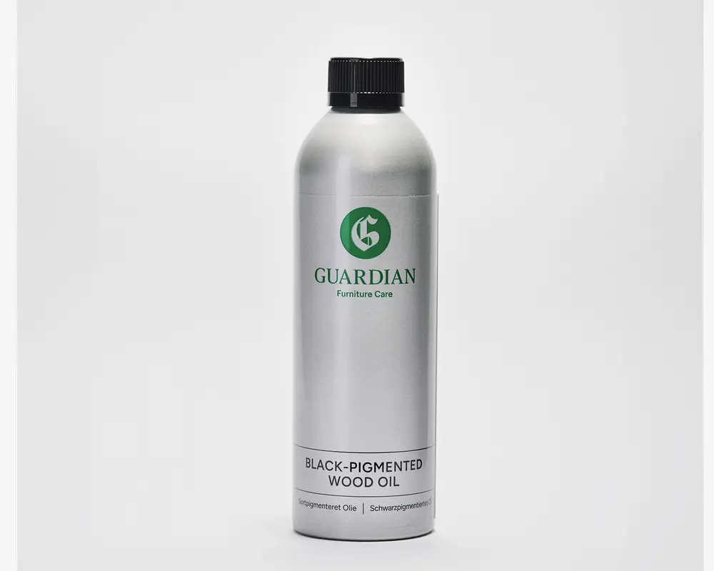 En høj, cylindrisk sølvfarvet metalflaske med sort skruelåg. Etiketten har mærket 'Guardian Furniture Care' i grøn samt et grønt cirkulært logo. Nederst på etiketten står der 'Black-Pigmented Wood Oil' med fed sort skrift, og der er yderligere tekst i mindre skrift nedenunder. Designet er minimalistisk og professionelt.