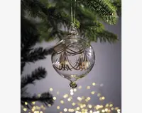 En klar glaskugle med en rund, let aflang form, dekoreret med fine guldfarvede mønstre og indgraverede stjernedesigns. Kuglen har en lille glasperle i bunden og hænger i en gylden snor. Den er ophængt på en grøn grangren, med bløde, varme lys sløret i baggrunden.