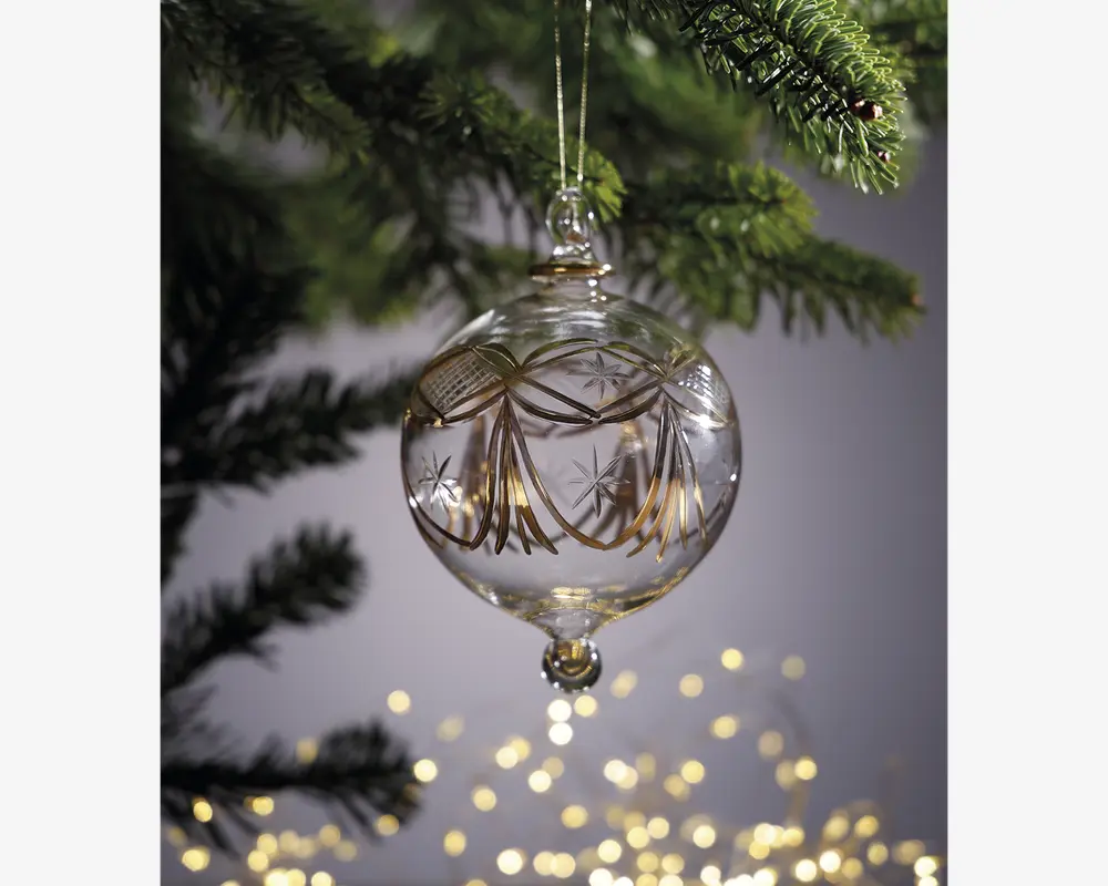 En klar glaskugle med en rund, let aflang form, dekoreret med fine guldfarvede mønstre og indgraverede stjernedesigns. Kuglen har en lille glasperle i bunden og hænger i en gylden snor. Den er ophængt på en grøn grangren, med bløde, varme lys sløret i baggrunden.