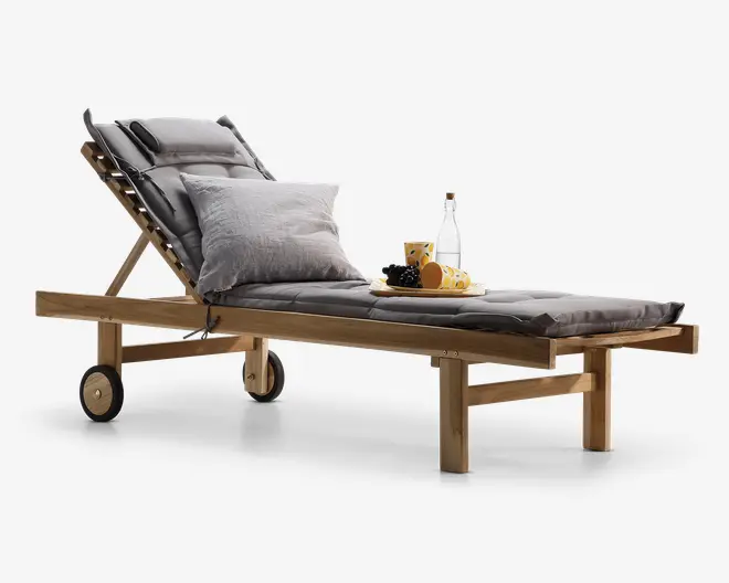 Justerbar liggestol i træ med en tyk, mørkegrå hynde og matchende nakkestøtte. Stolen har et ryglæn, der kan lænes tilbage, to solide hjul for nem flytning og et bredt sæde. Den er udstyret med en stor, lysegrå pude og en bakke med en flaske, en kop og en tallerken med frugt, hvilket giver et indtryk af komfort og afslapning.