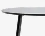 Nærbillede af en rund bordplade med en glat, lys overflade og en tynd, mørk kant. Bordet har et solidt, cylindrisk ben med mat sort finish, som er let vinklet udad for stabilitet. Designet er moderne og minimalistisk med fokus på rene linjer og enkel konstruktion.