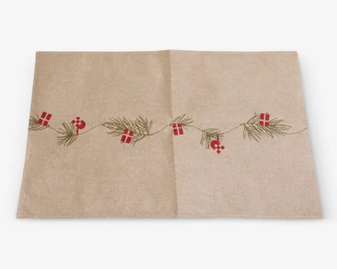 Rektangulært beige dækkeserviet med et festligt broderet design langs midten. Broderiet består af grønne grangrene, små røde gaveæsker med hvide bånd og røde juleornamenter, hvilket giver et julet udtryk. Stoffet har en let tekstur, og dækkeservietten har pæne, lige kanter.