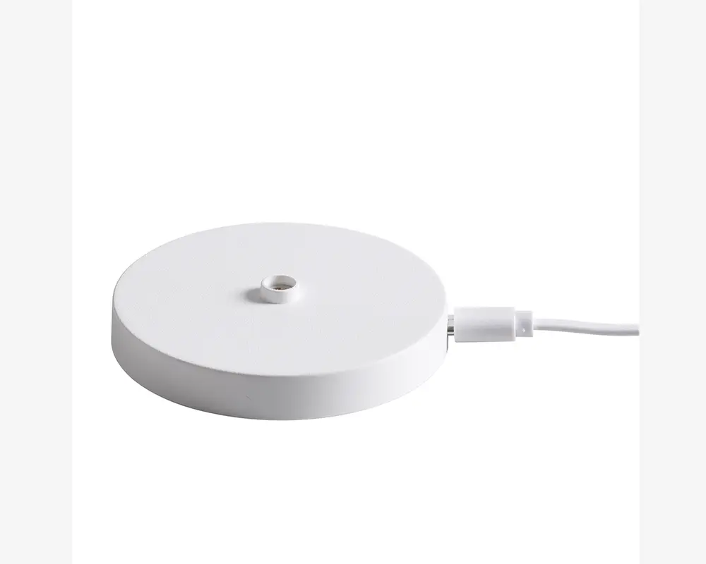 En rund, flad, hvid base med en glat, mat overflade. Der er et lille, centreret hul på toppen, sandsynligvis til montering af en anden komponent. En hvid strømkabel er tilsluttet siden, hvilket indikerer, at basen er strømforsynet. Designet er minimalistisk og moderne.